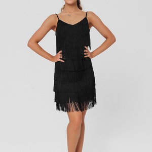 May include: A woman wearing a black fringe dress and brown heeled sandals. She is standing with her hands on her hips and smiling at the camera.