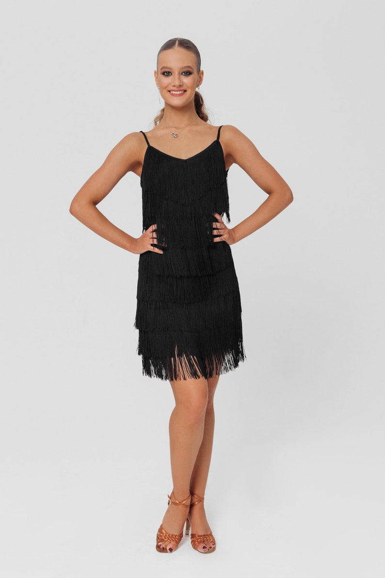 May include: A woman wearing a black fringe dress and brown heeled sandals. She is standing with her hands on her hips and smiling at the camera.