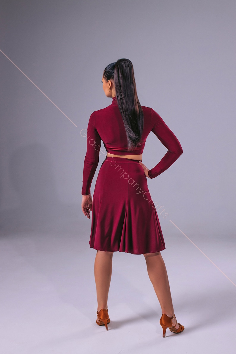 May include: A woman wearing a maroon long-sleeved crop top and a matching A-line skirt. She is wearing brown heeled shoes and has her hair pulled back in a ponytail.