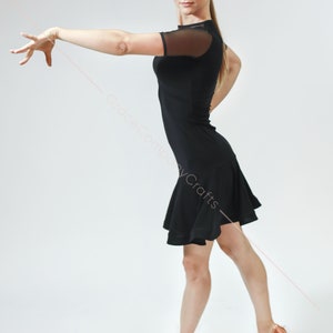 May include: A woman wearing a black dance dress with a mesh overlay on the sleeves. She is posing with her right arm extended and her left hand on her hip. She is wearing brown dance shoes.