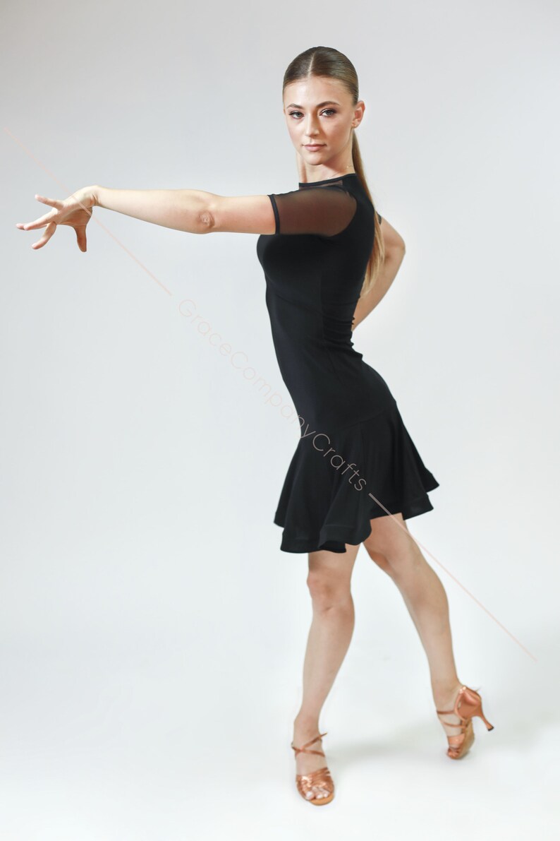 May include: A woman wearing a black dance dress with a mesh overlay on the sleeves. She is posing with her right arm extended and her left hand on her hip. She is wearing brown dance shoes.