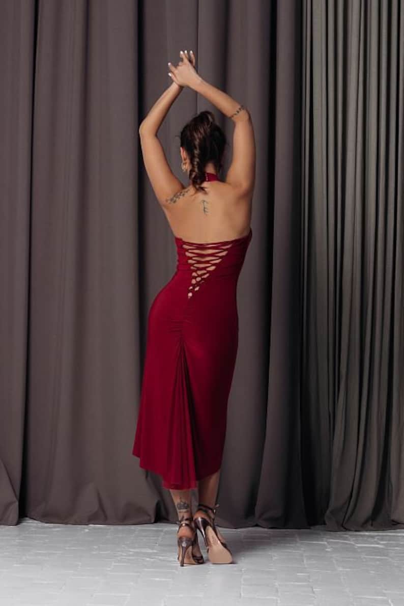 May include: A woman wearing a red halter dress with a criss-cross back detail. The dress is fitted and has a flowing skirt. She is wearing high heels and has her arms raised above her head.