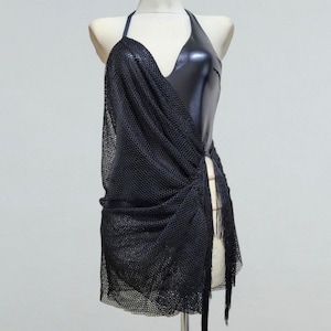 May include: A black halter dress on a mannequin. The dress features a shiny, dark bodice and a draped, mesh overlay. The overlay is gathered at the waist and has fringe detailing along the side. The dress is designed for a sleek, modern look.