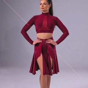 May include: A woman wearing a maroon dance outfit with a long-sleeved crop top and a fringed skirt. She is wearing brown dance shoes.