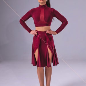 May include: A woman wearing a maroon dance outfit with a long-sleeved crop top and a skirt with fringe accents and side slits. She is wearing brown dance shoes.