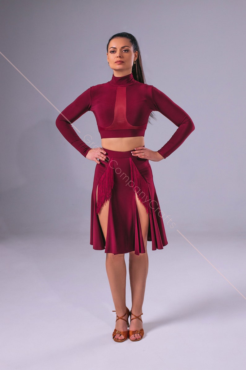 May include: A woman wearing a maroon dance outfit with a long-sleeved crop top and a skirt with fringe accents and side slits. She is wearing brown dance shoes.