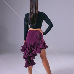 May include: A woman wearing a black long-sleeved crop top and a purple ruffled skirt. She is standing with her back to the camera and her right hand on her hip. She is wearing brown heeled shoes.
