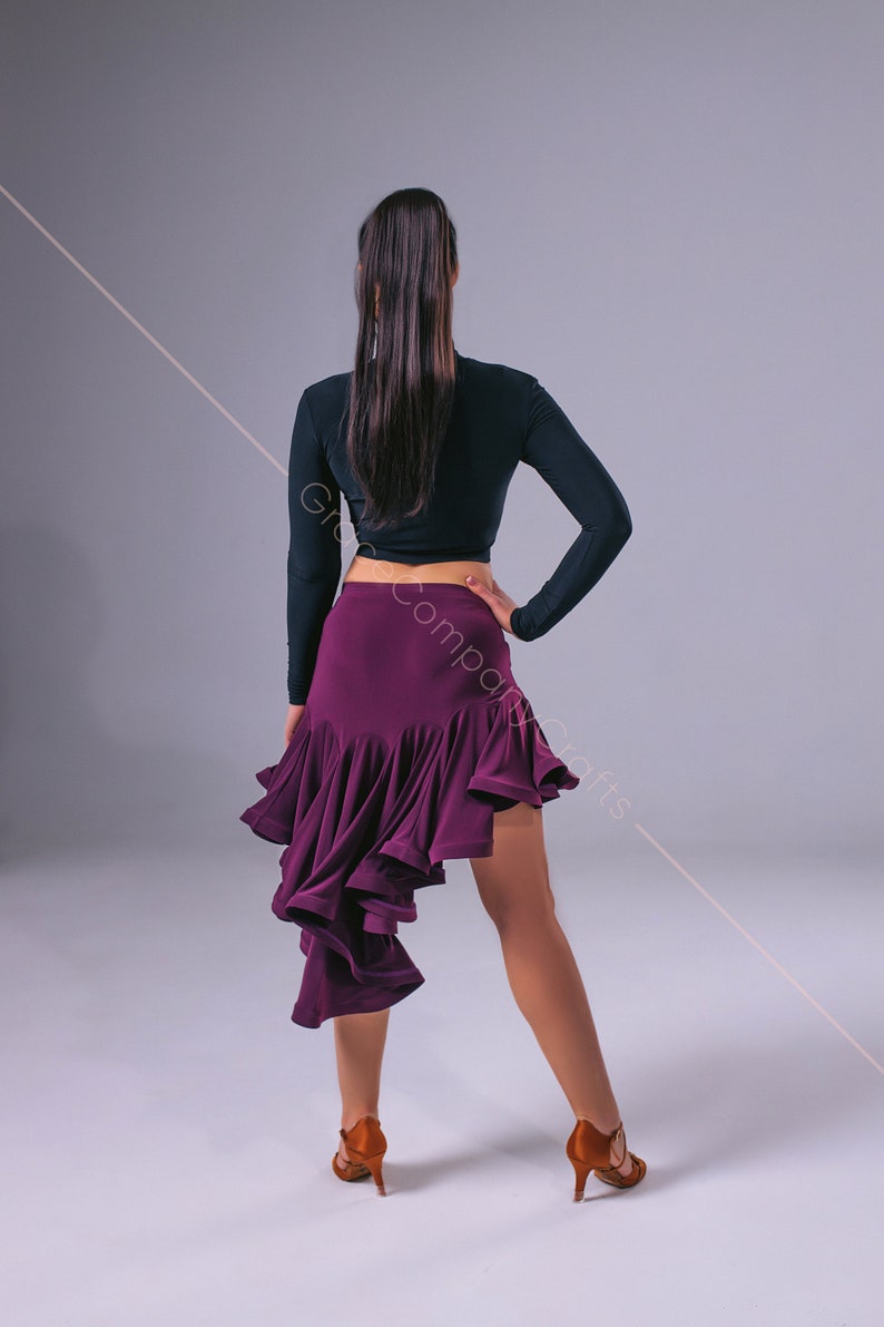 May include: A woman wearing a black long-sleeved crop top and a purple ruffled skirt. She is standing with her back to the camera and her right hand on her hip. She is wearing brown heeled shoes.