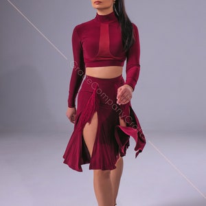 May include: A woman wearing a burgundy long-sleeved crop top and a matching skirt with a front slit and fringe detail. She is wearing brown heeled sandals and has her long dark hair pulled back in a ponytail.
