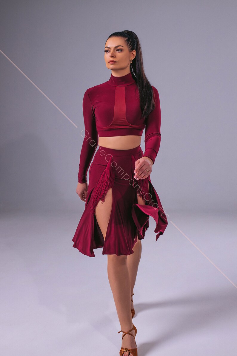 May include: A woman wearing a burgundy long-sleeved crop top and a matching skirt with a front slit and fringe detail. She is wearing brown heeled sandals and has her long dark hair pulled back in a ponytail.