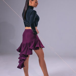 May include: A woman wearing a black long-sleeved top and a purple ruffled skirt. She is wearing brown dance shoes and has her hair pulled back in a ponytail.
