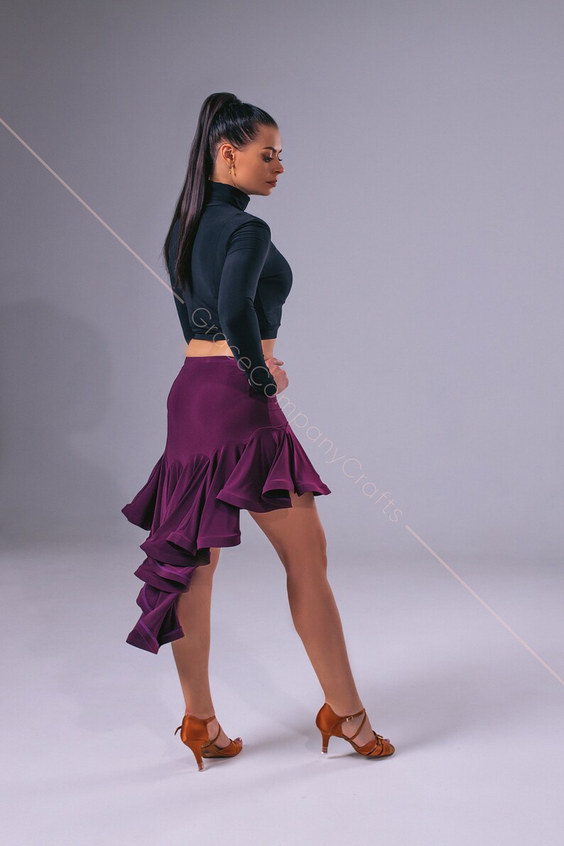 May include: A woman wearing a black long-sleeved top and a purple ruffled skirt. She is wearing brown dance shoes and has her hair pulled back in a ponytail.