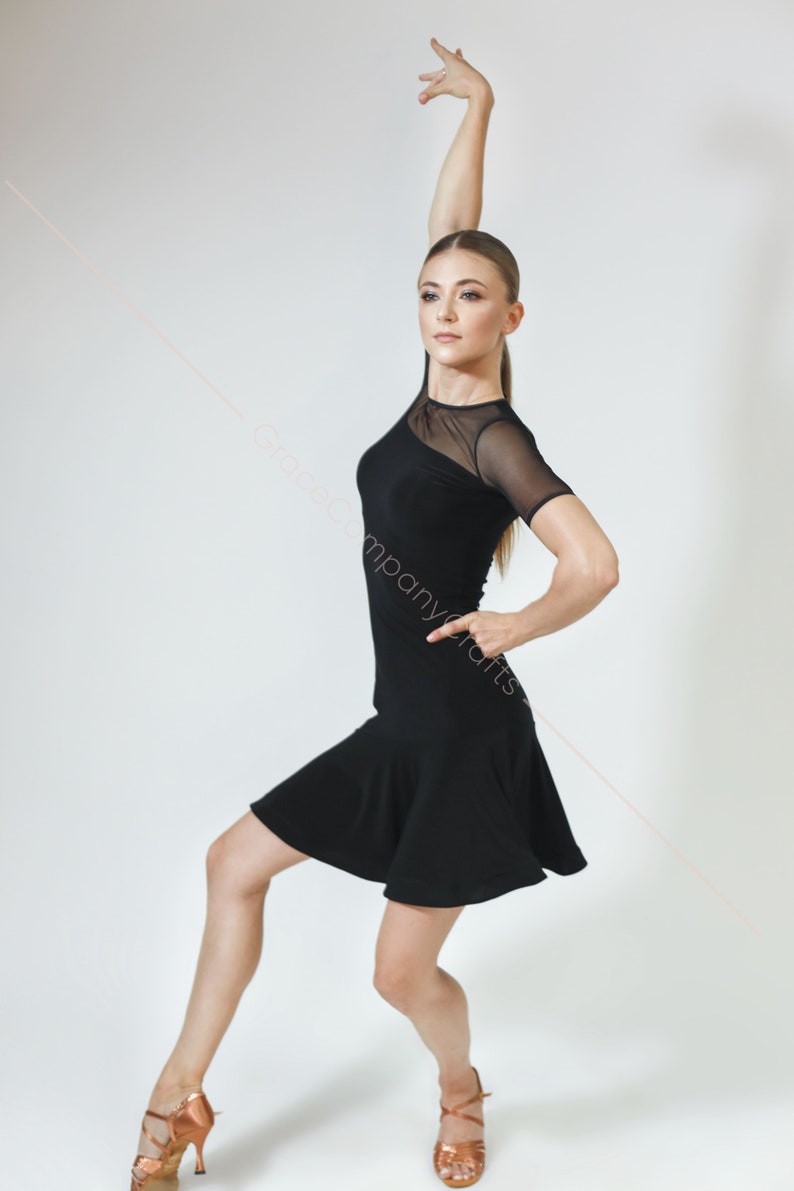 May include: A woman in a black dance dress with a sheer mesh overlay and a flared skirt. She is wearing brown dance shoes and is posing with her arm raised.