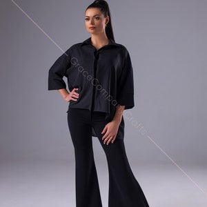Puede incluir: Una mujer con una camisa negra de gran tama&ntilde;o con cuello y pantalones acampanados negros. Est&aacute; de pie sobre un fondo gris.