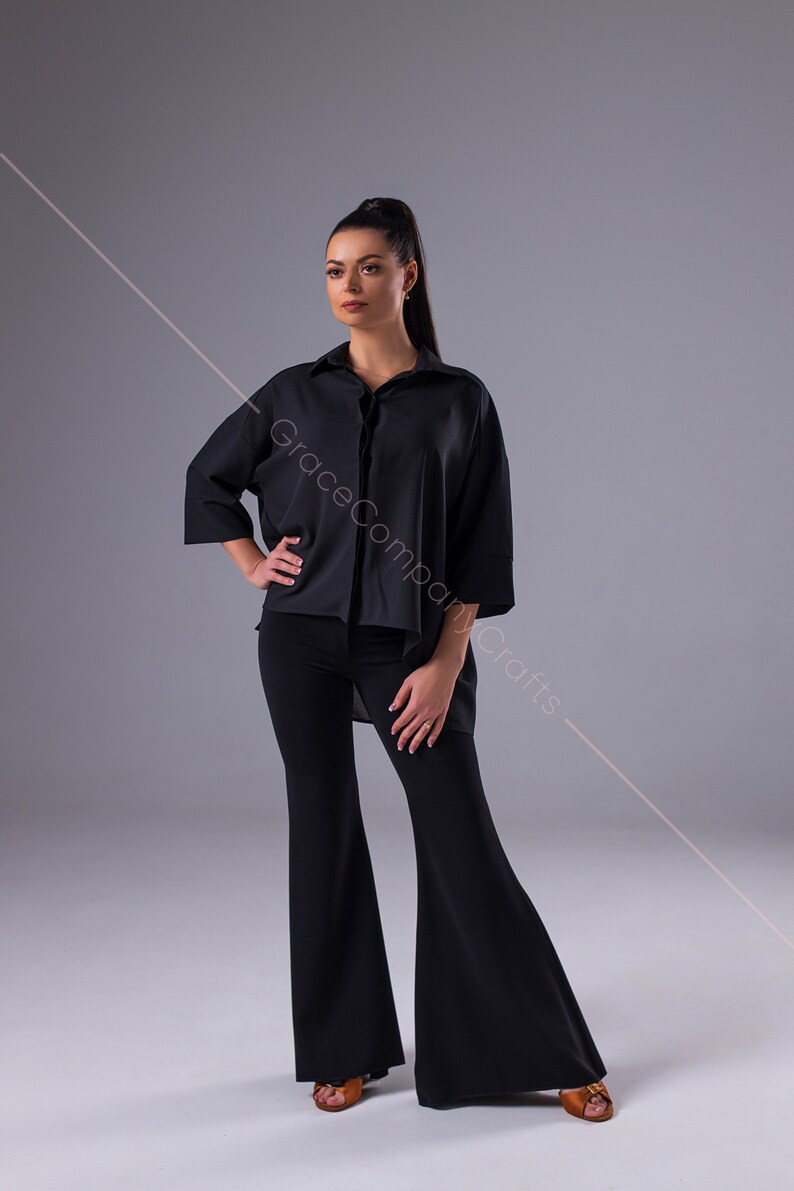 Puede incluir: Una mujer con una camisa negra de gran tama&ntilde;o con cuello y pantalones acampanados negros. Est&aacute; de pie sobre un fondo gris.