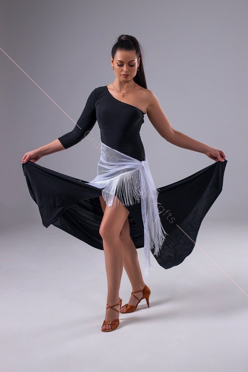 May include: A woman wearing a black one-shoulder dress with a white fringed shawl and brown dance shoes. She is posing with her arms outstretched and the skirt of her dress flowing behind her.
