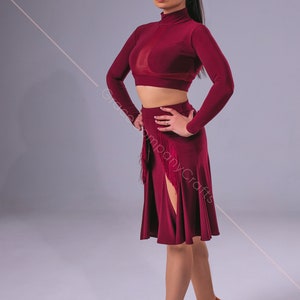 May include: A woman wearing a maroon long-sleeved crop top and a maroon skirt with a side slit and fringe detail. She is wearing brown heeled sandals.