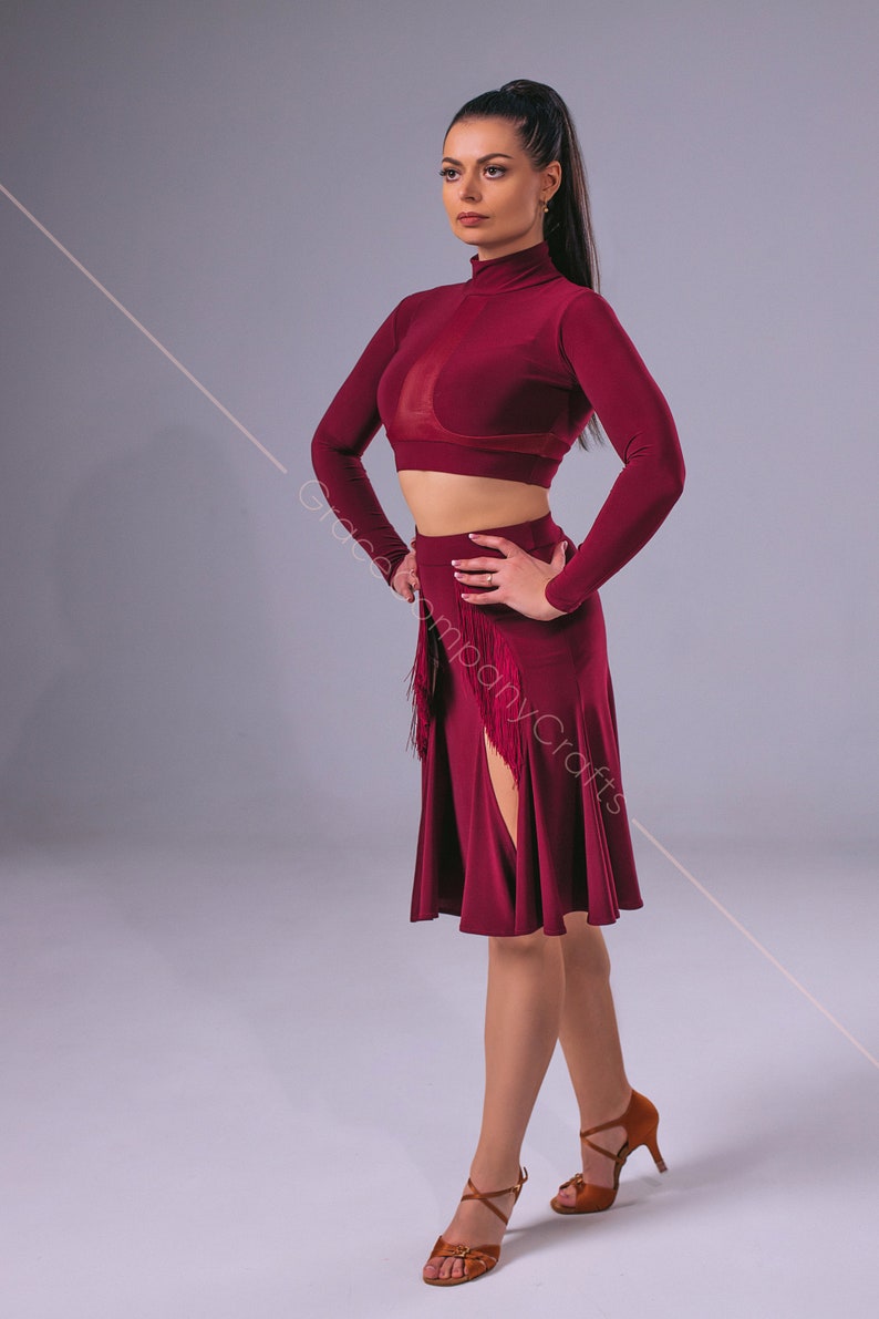 May include: A woman wearing a maroon long-sleeved crop top and a maroon skirt with a side slit and fringe detail. She is wearing brown heeled sandals.