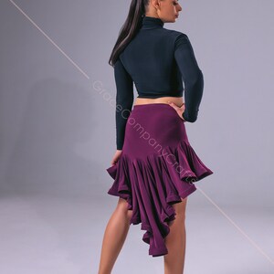 May include: A woman wearing a black long-sleeved crop top and a purple asymmetrical dance skirt. She is wearing brown dance shoes and has her hair pulled back in a ponytail.