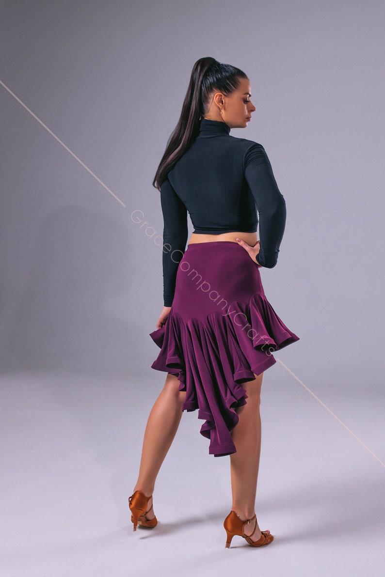 May include: A woman wearing a black long-sleeved crop top and a purple asymmetrical dance skirt. She is wearing brown dance shoes and has her hair pulled back in a ponytail.
