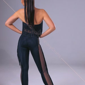 May include: A black dance outfit with fringe detailing. The outfit includes a strapless top and leggings with a sheer panel on one leg. The leggings have fringe trim along the sides. The model is wearing brown heels.