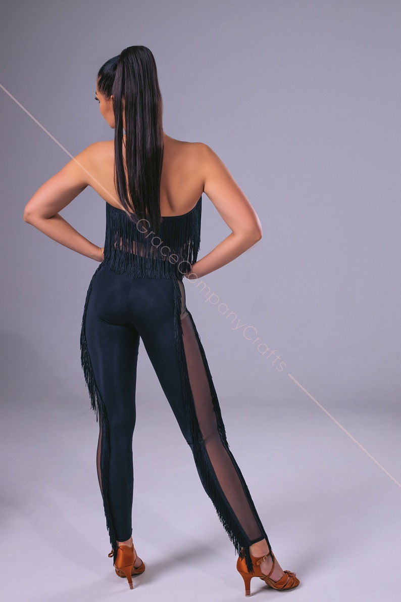 May include: A black dance outfit with fringe detailing. The outfit includes a strapless top and leggings with a sheer panel on one leg. The leggings have fringe trim along the sides. The model is wearing brown heels.