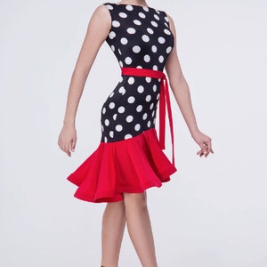 Op de afbeelding: Een zwart-witte polkadotjurk met een rode geruite rok en een rode sash. De jurk is mouwloos en heeft een ronde hals.