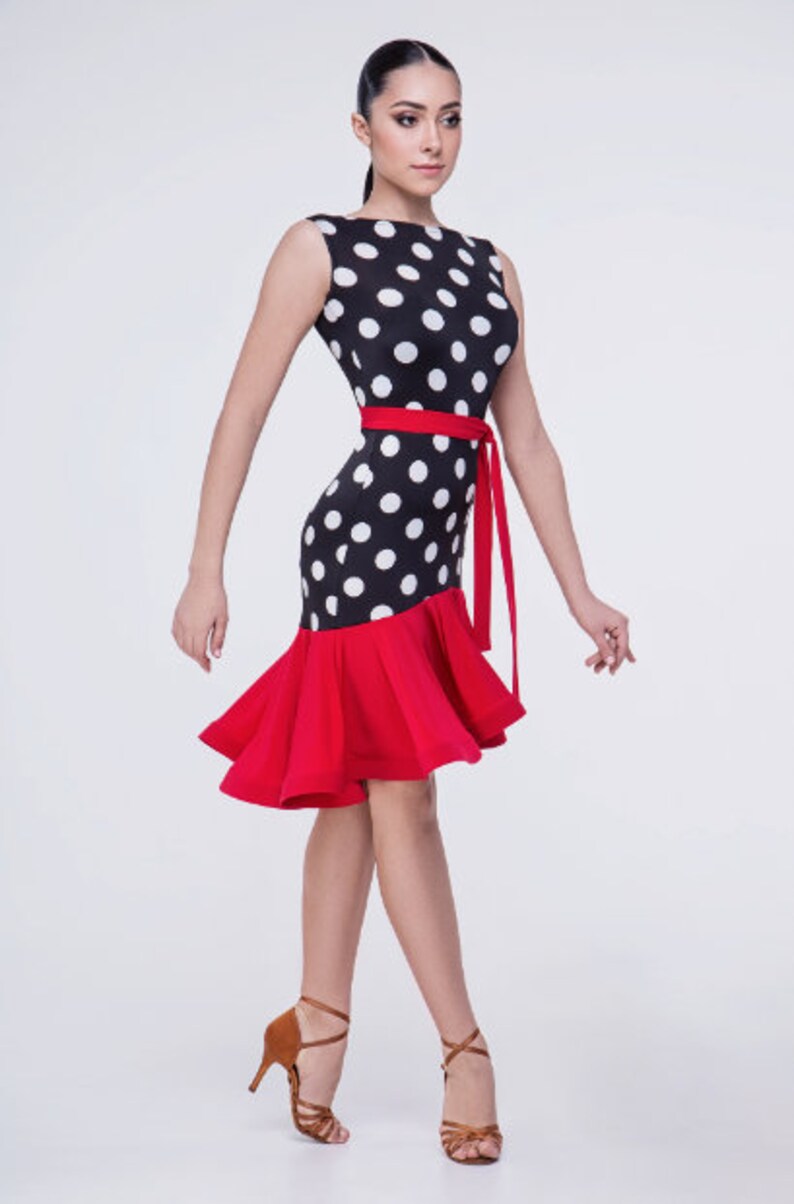 Op de afbeelding: Een zwart-witte polkadotjurk met een rode geruite rok en een rode sash. De jurk is mouwloos en heeft een ronde hals.