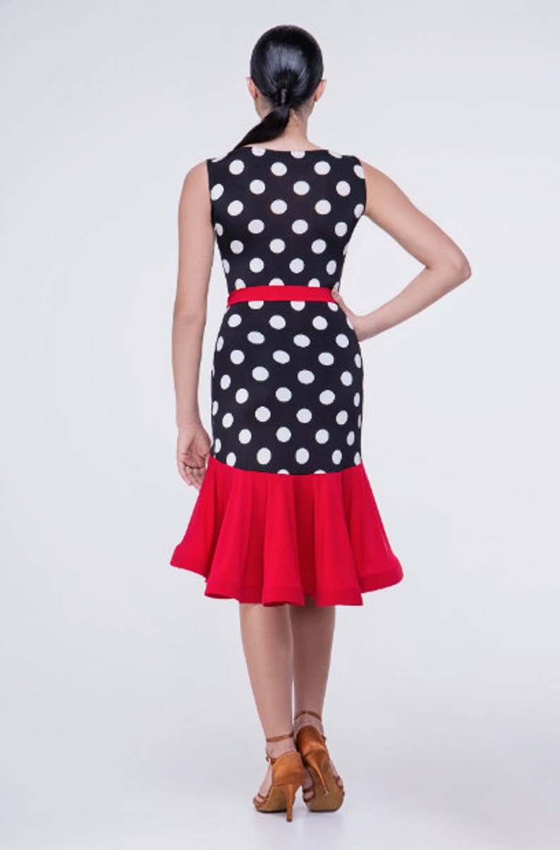 Op de afbeelding: Een zwart-witte polkadotjurk met een rode geruite zoom en een rode riem. De jurk is mouwloos en heeft een ronde hals.