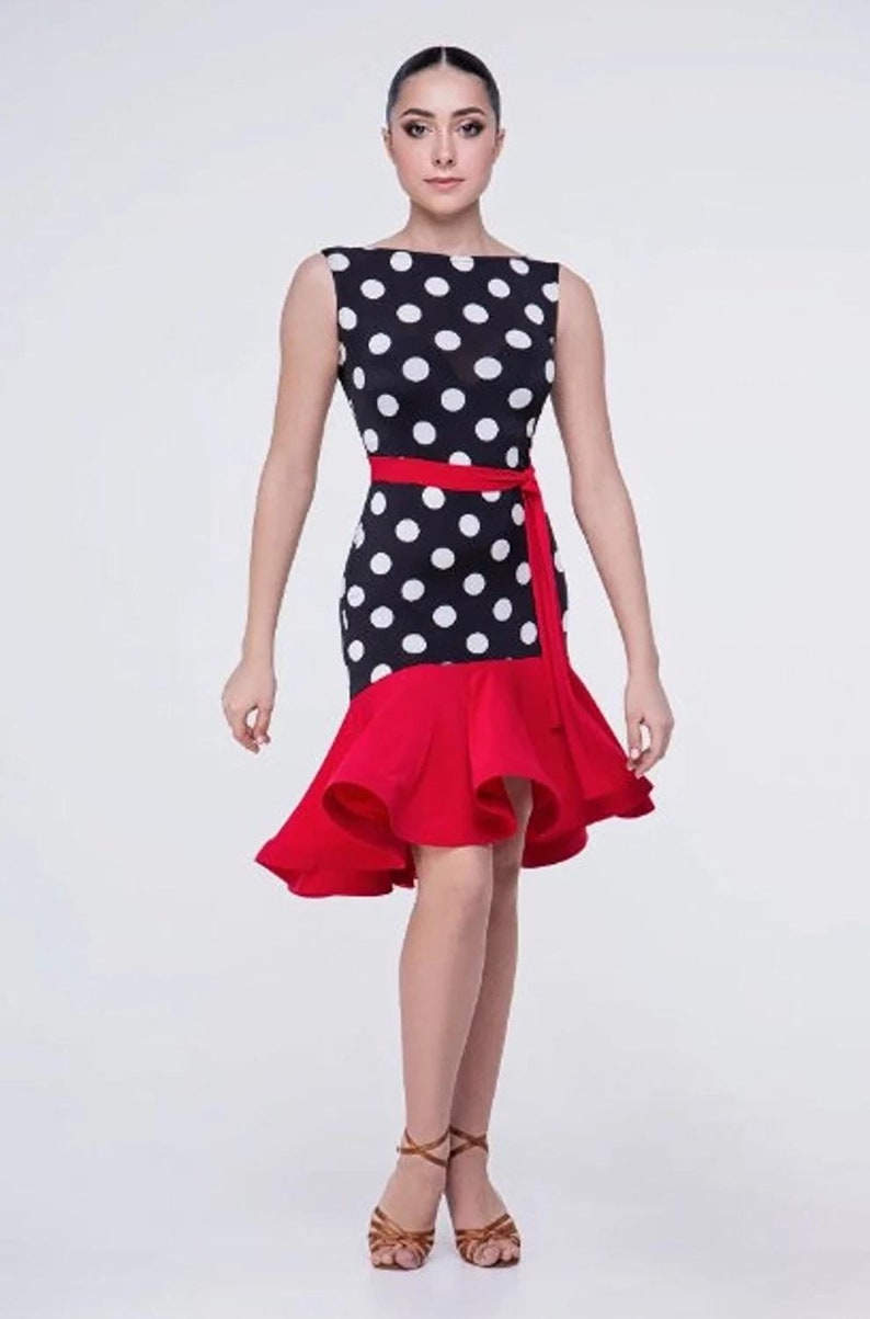 Op de afbeelding: Een zwart-witte polkadotjurk met een rode geruite zoom en een rode sash. De jurk is mouwloos en heeft een ronde hals.