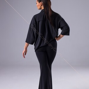 Puede incluir: Una mujer con una camisa negra de manga larga con cuello y pantalones negros acampanados. La camisa tiene un corte holgado y los pantalones son ajustados en las caderas y los muslos.