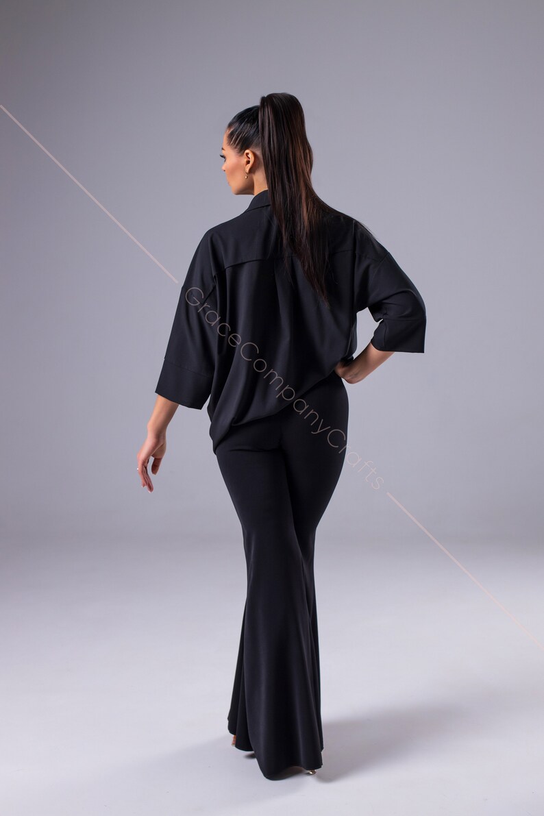Puede incluir: Una mujer con una camisa negra de manga larga con cuello y pantalones negros acampanados. La camisa tiene un corte holgado y los pantalones son ajustados en las caderas y los muslos.