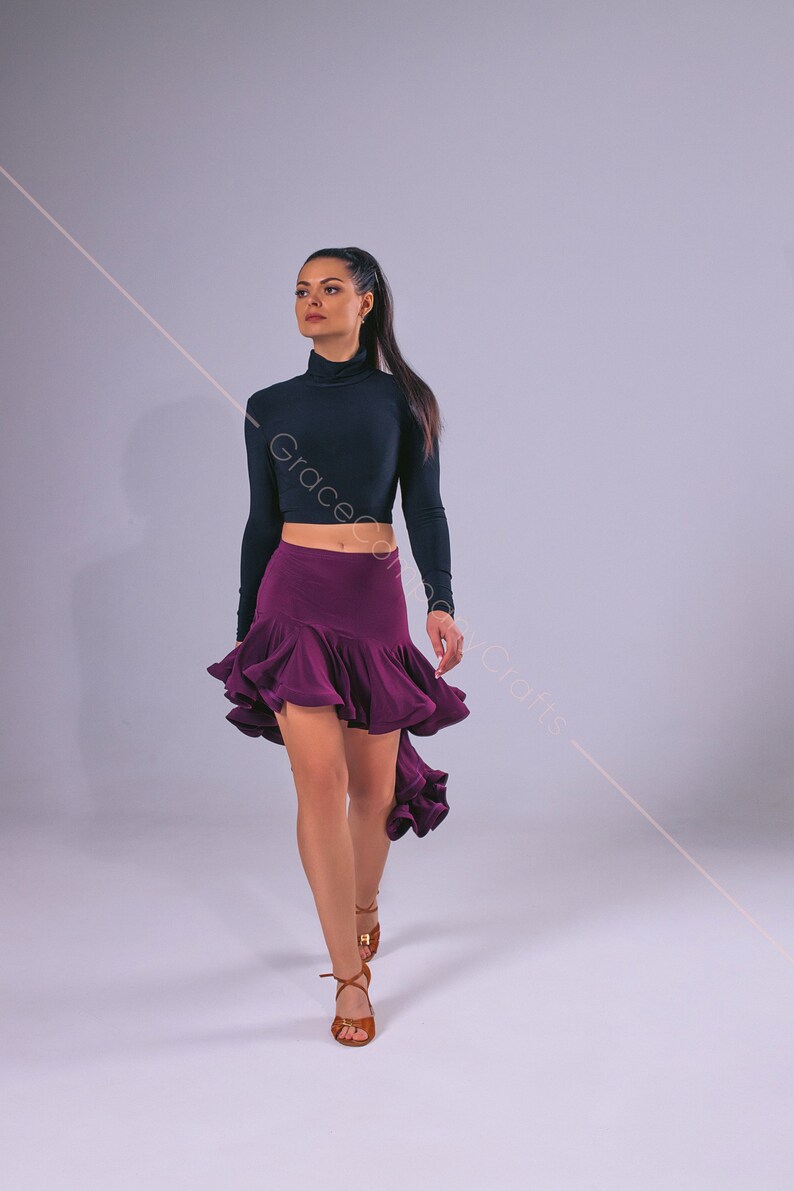 May include: A woman wearing a black long-sleeved crop top and a purple ruffled skirt. She is wearing brown dance shoes and is walking towards the camera.