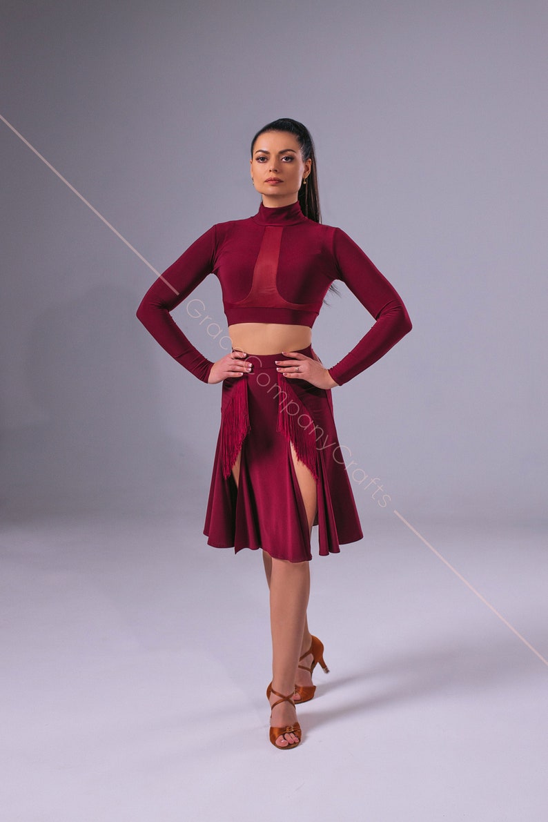 May include: A woman wearing a burgundy dance outfit with a long-sleeved crop top and a skirt with a slit and fringe detail. She is wearing brown dance shoes.
