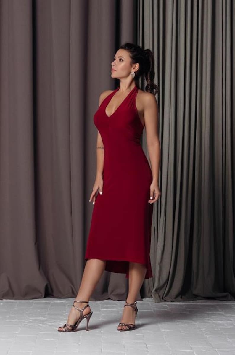 May include: A woman wearing a red sleeveless dress with a high neckline and a high slit. She is standing in front of a grey curtain and wearing silver heels.