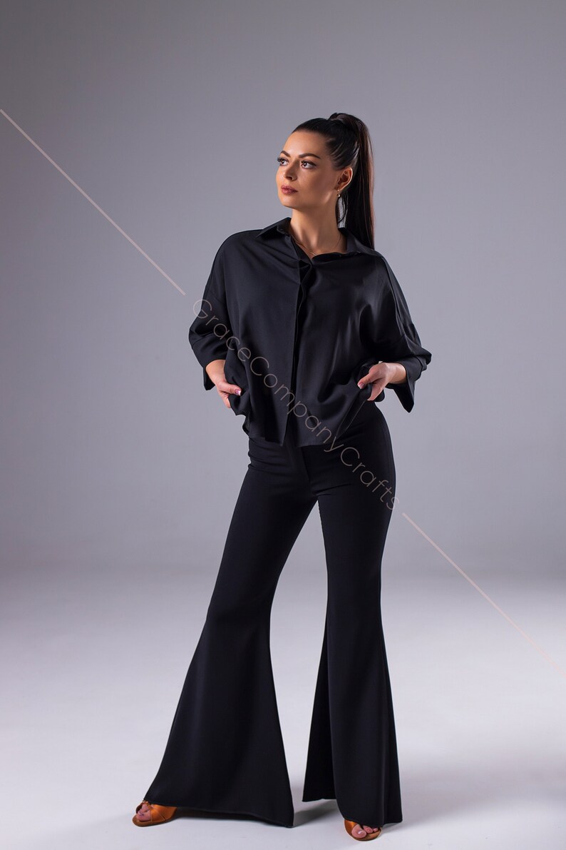 Puede incluir: Una mujer con una camisa negra de cuello y pantalones acampanados negros. Est&aacute; de pie con las manos en las caderas y mirando hacia un lado.