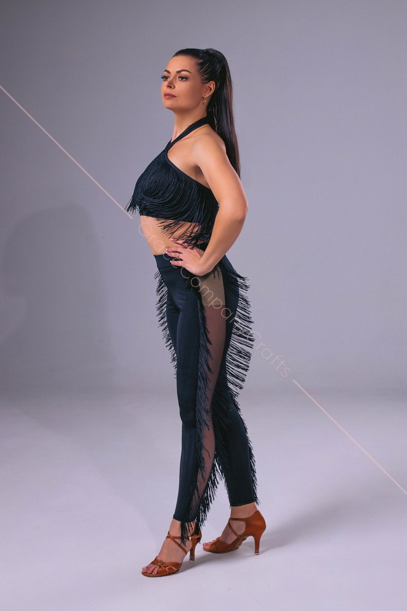 May include: A woman wearing a black dance outfit with fringe details. The outfit includes a halter top and leggings. She is wearing brown dance shoes.