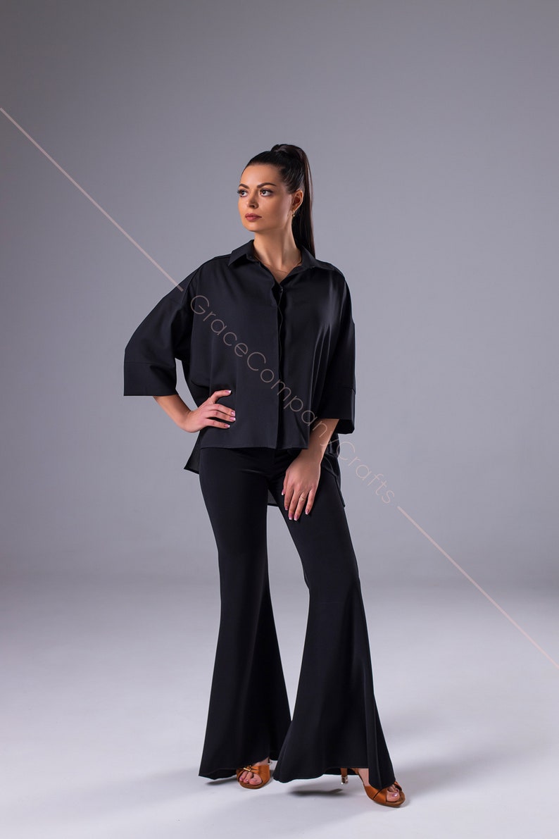 Puede incluir: Una mujer con una camisa negra de manga larga con cuello y pantalones negros acampanados. Est&aacute; de pie sobre un fondo gris.