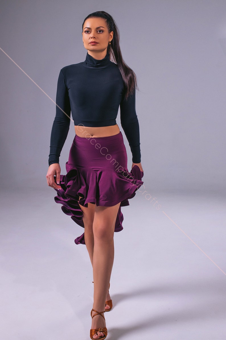 May include: A woman wearing a black long-sleeved crop top and a purple ruffled skirt. She is wearing brown dance shoes and has her hair pulled back in a ponytail.