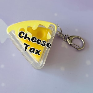May include: A yellow cheese wedge keychain with the text "Cheese Tax" in black. The keychain has a clear plastic border and a silver-colored clasp. The background is a light purple color with white star shapes.