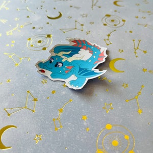May include: A blue and red dragon sticker with a white background. The sticker is on a light blue background with gold foil stars, moons, and constellations.