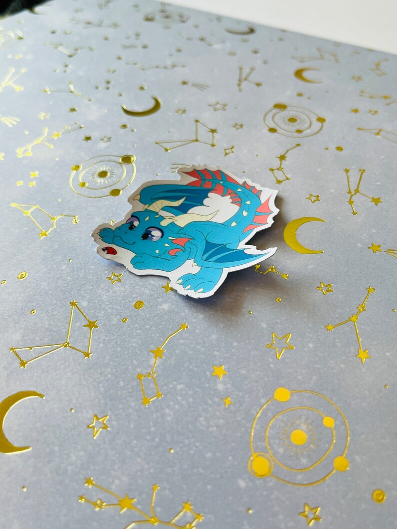 May include: A blue and red dragon sticker with a white background. The sticker is on a light blue background with gold foil stars, moons, and constellations.
