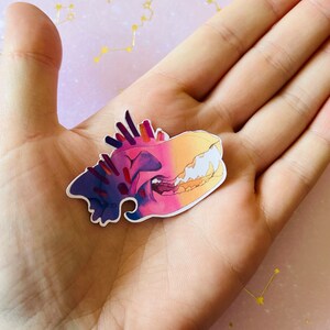 May include: A colorful sticker of a skull with a gradient of purple, pink, and yellow. The skull has sharp teeth and a detailed design.