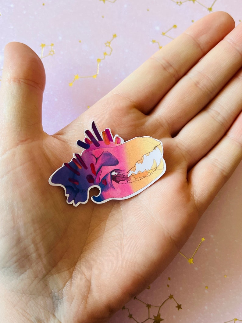 May include: A colorful sticker of a skull with a gradient of purple, pink, and yellow. The skull has sharp teeth and a detailed design.