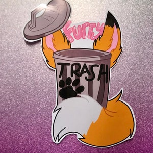 May include: A gray trash can with a black paw print and the word "TRASH" written on it. The trash can is surrounded by a cartoon fox's ears and tail. The word "Furry" is written above the trash can.