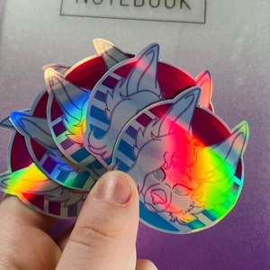 May include: A set of four iridescent holographic stickers featuring a cartoon animal with blue and pink accents. The stickers are round and have a white background with a blue and pink striped pattern.