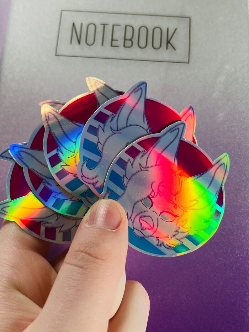 May include: A set of four iridescent holographic stickers featuring a cartoon animal with blue and pink accents. The stickers are round and have a white background with a blue and pink striped pattern.