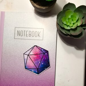 May include: A galaxy-themed sticker of a 20-sided die, or d20, on a silver and purple notebook with the word "NOTEBOOK" printed on the front.
