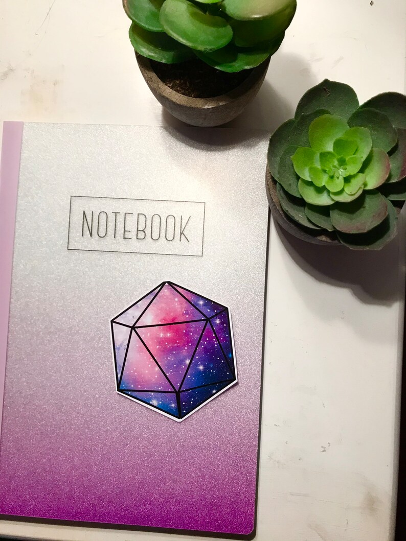 May include: A galaxy-themed sticker of a 20-sided die, or d20, on a silver and purple notebook with the word "NOTEBOOK" printed on the front.