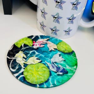 May include: A circular coaster with a koi pond design, including fish, lily pads, and water effects. Beside it is a white mug with a repeating pattern of a bird wearing sunglasses and a star.